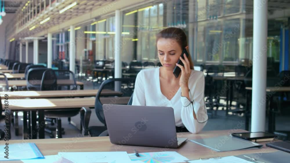 businesswoman typing on laptop talking by mobile with client young caucasian manager working in the open space office. woman with blond hair wearing casual white shirt sitting at the workplace businesswoman typing on laptop talking by mobile with client young caucasian manager working in the open space office. woman with blond hair wearing casual white shirt sitting at the workplace