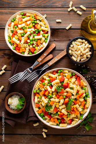 Italian pasta salad with fusilli, corn, boiled carrot, fresh green peas and sweet pepper on wooden rustic background. Top view