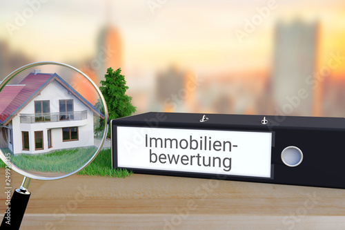 Wert einer Immobilie ermitteln. Ordner beschriftet mit dem Begriff Immobilienbewertung liegt neben einem Haus-Modell mit Lupe auf einem Schreibtisch. Skyline einer Stadt im Hintergrund.