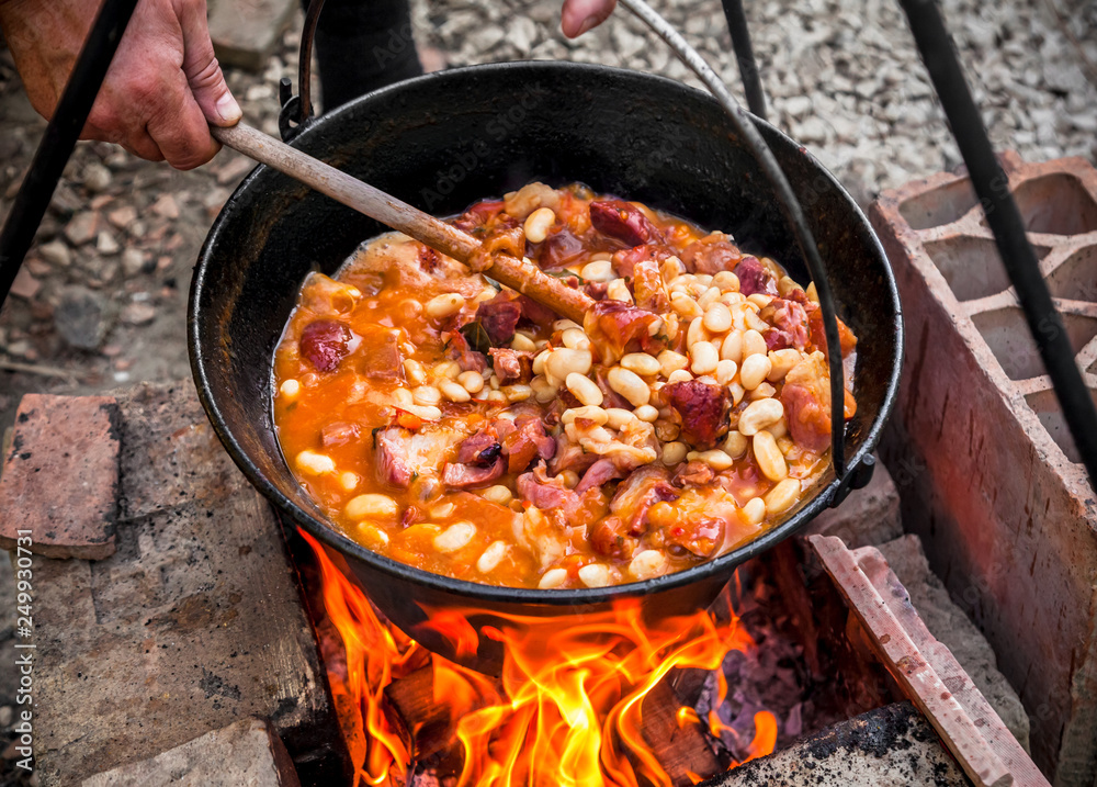 Cooking beans with bacon and sausages outdoors in black pot at picnic party on fire Cooking beans with bacon and sausages outdoors in black pot at picnic party on fire