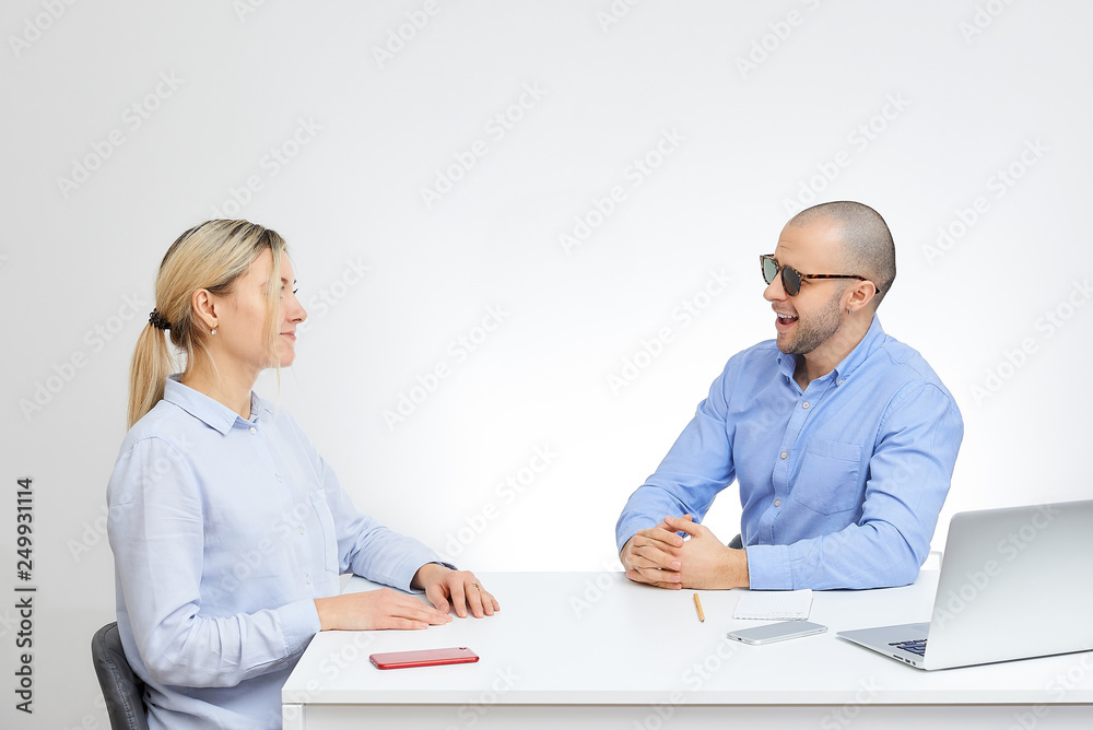 Happy brutal boss in a blue shirt with glasses interviews new blonde caucasian employee sits by the desk in the office with the white background. Happy brutal boss in a blue shirt with glasses interviews new blonde caucasian employee sits by the desk in the office with the white background.
