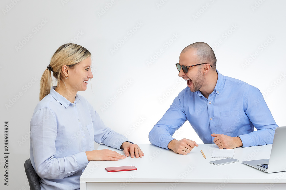 Laughing brutal boss in sunglasses in a blue shirt jokes with his blonde caucasian colleague sits by the desk in the office with the white background. Laughing brutal boss in sunglasses in a blue shirt jokes with his blonde caucasian colleague sits by the desk in the office with the white background.