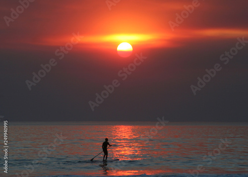 Male paddle boarder moves across ocean in front of amazing red sunset.