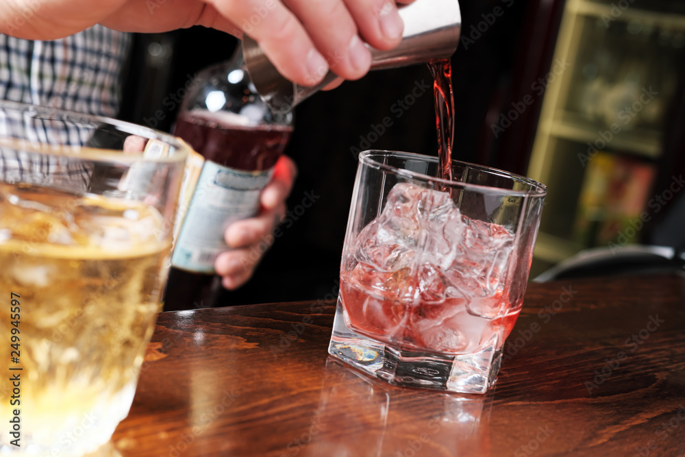 Close up shot of bartender hand pouring drink from measuring cup into a cocktail glass filled with ice cubes. Barman preparing fresh negroni cocktail Close up shot of bartender hand pouring drink from measuring cup into a cocktail glass filled with ice cubes. Barman preparing fresh negroni cocktail