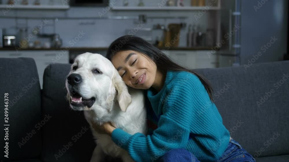 Atractive indian woman sitting with dog on couch at home. Loving young owner smiling, looking at camera while embracing and stroking her pet golden retriever on sofa in domestic room. Atractive indian woman sitting with dog on couch at home. Loving young owner smiling, looking at camera while embracing and stroking her pet golden retriever on sofa in domestic room.