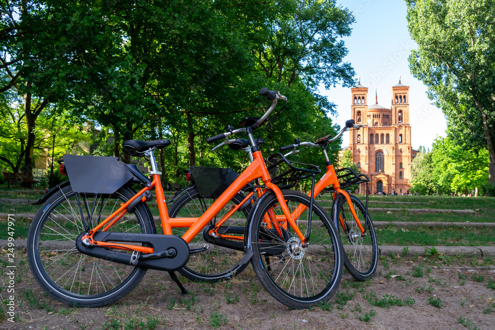Two orange bicycles parked towards St. Thomas protestant church (Thomaskirche) in Berlin, Germany. Urban bike share concept for weekend gateaways and sightseeing eco friendly city cycle tours.