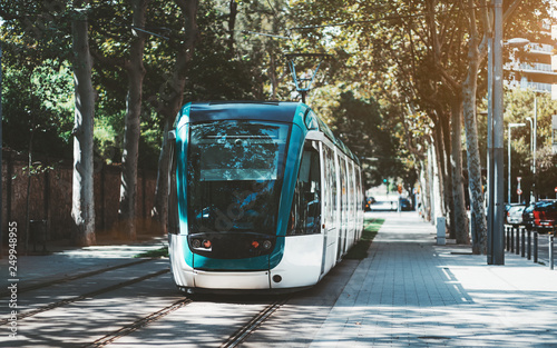 A modern neat tram in city alleyway surrounded by trees; blue and white city streetcar near the sidewalk made of pavement stones; contemporary tramcar on the station with the road on the right photo