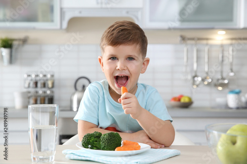 Adorable little boy eating vegetables at table in kitchen