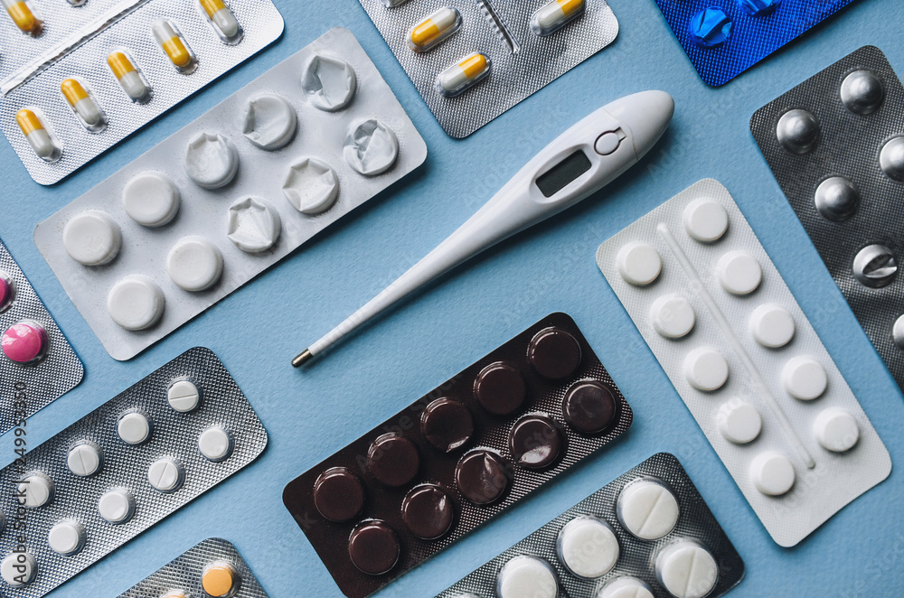 A variety of colored pills and white electronic thermometer lie on a blue background. Modern medicine and drugs. Flat lay, top view. A variety of colored pills and white electronic thermometer lie on a blue background. Modern medicine and drugs. Flat lay, top view.