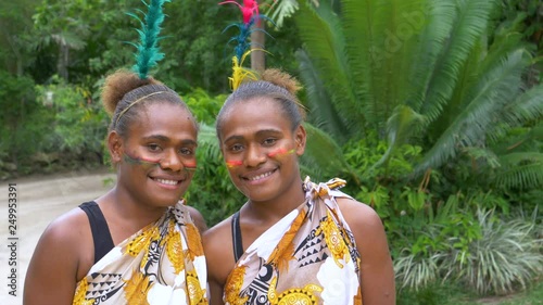 SLOW MOTION, PORTRAIT: Beautiful Melanesian sisters dressed into traditional outfits stand together and smile at camera. Lovely friends wearing vibrant indigenous make up and feathers in their hair.