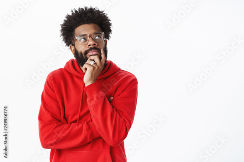 Creative african american bearded guy with afro hairstyle in glasses and red hoodie creating new song, standing in thoughtful pose touching chin looking dreamy, focused at upper right corner, thinking