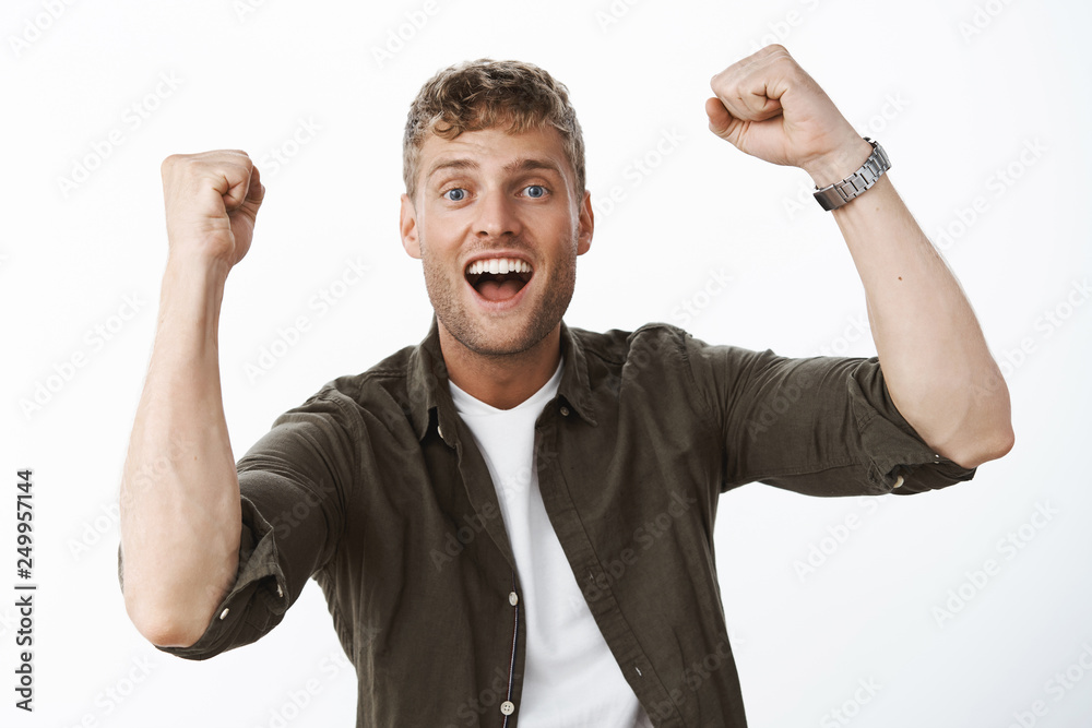 Fototapeta premium Optimistic and excited charismatic blond guy with blue eyes and white broad smile raising clenched fists in triumph, support and cheer smiling delighted from success and victory over gray wall
