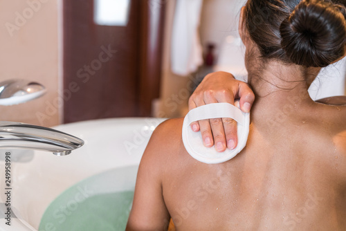 Obraz na plátně Woman taking a bath at luxury hotel scrubbing back skin with hand scrub sponge removing dead skin cells for body, skincare exfoliation