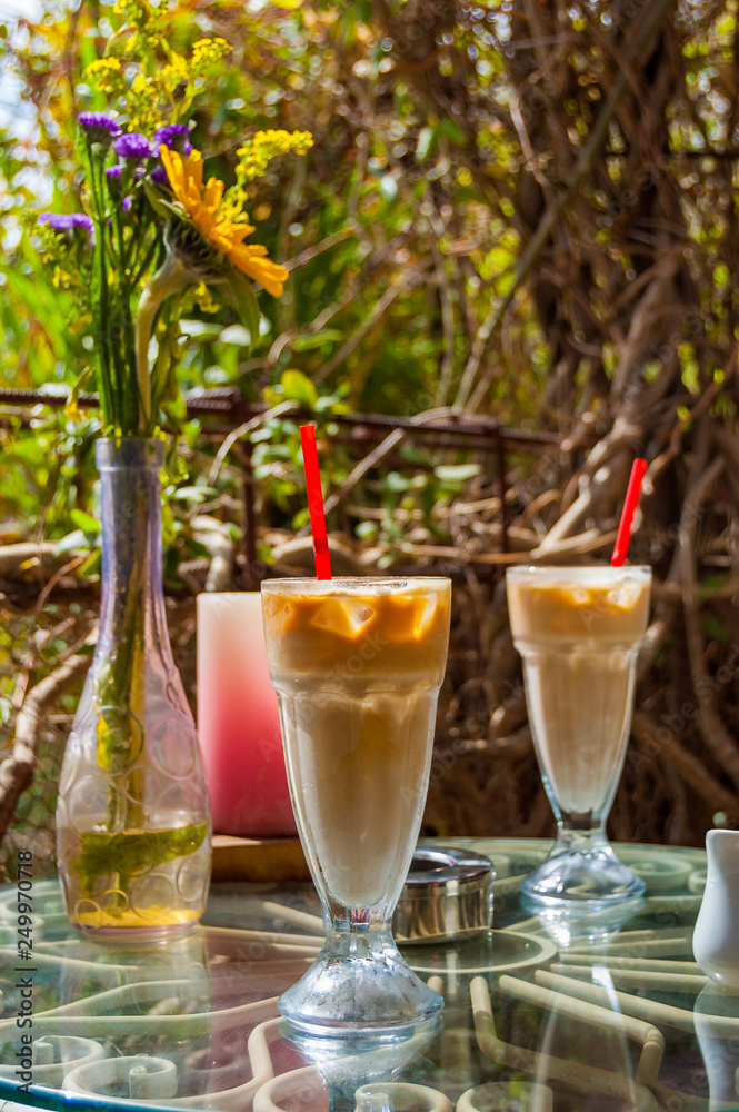 Two Ice coffee glasses with red drinking straw, vase with flowers, ashtray and red candle on the metal glass table surrounded by exotic plants. Romantic place.