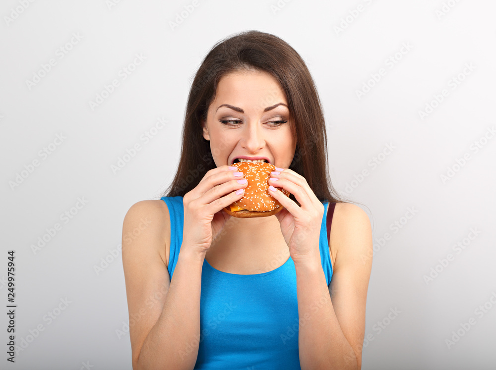 Thinking funny hungry excited woman eating burger and looking down on blue background.