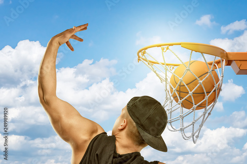 Man wearing black t-shirt and black cap throwing basketball ball into hoop on blue sky white clouds background