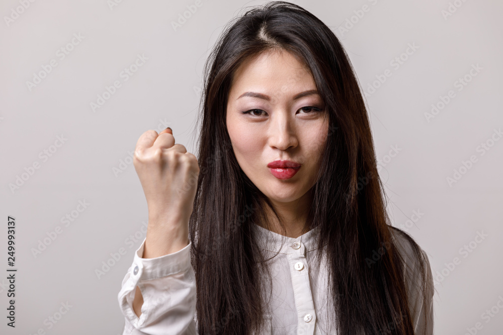 portrait of asian woman in white shirt showing fist on gray background. emotions portrait of asian woman in white shirt showing fist on gray background. emotions