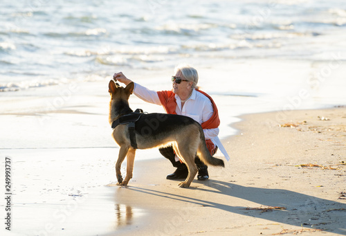 Happy attractive senior woman with her german shepard dog playing on the beach at autumn sunset