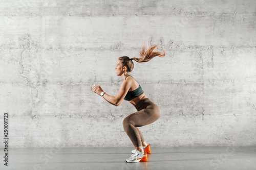 Side view of muscular focused brunette with ponytail and in sportswear jumping in front of brick wall in gym.