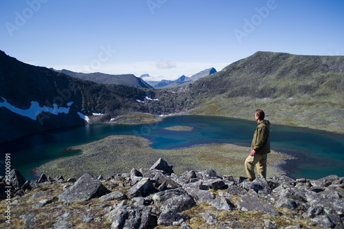A tourist in Hakka clothes stands on top of a mountain and looks at a blue lake, not far from the top of Narodnaya Mountain. Yugyd va National Park, Subpolar Urals, Komi Republic, Russia