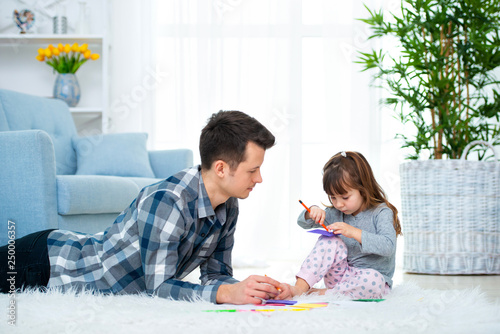 father and little daughter having quality family time together at home. dad with girl lying on warm floor drawing with colorful felt tip pencils.