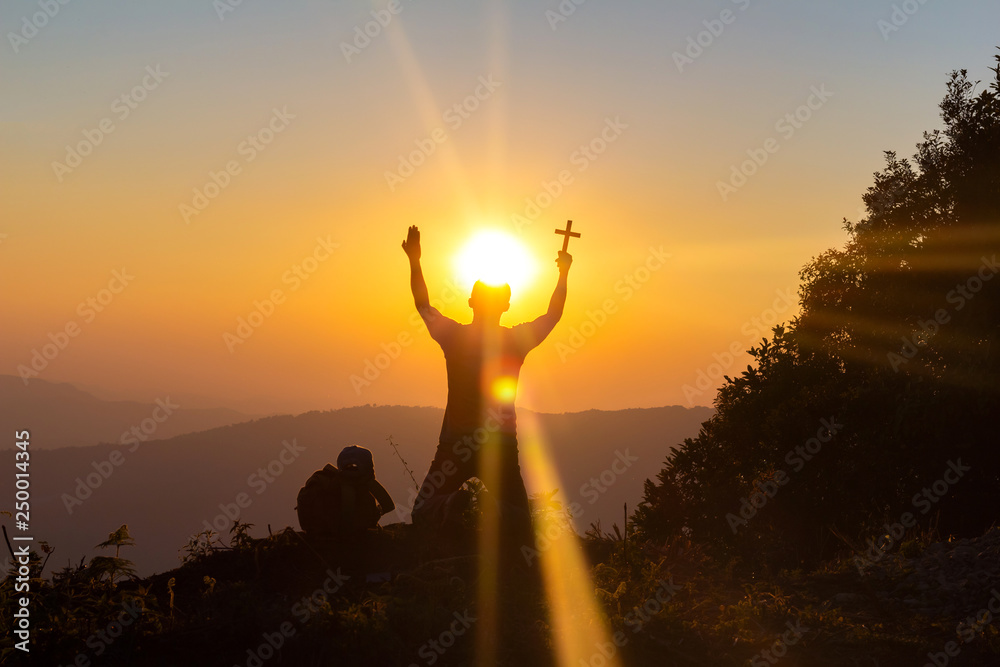 human praying to the GOD while holding a crucifix symbol with bright sunbeam on mountain at sunset time