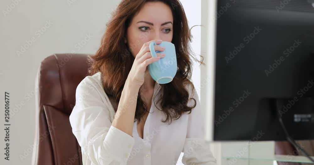 Beautiful businesswoman working in white office looking at computer monitor drinking coffee Beautiful businesswoman working in white office looking at computer monitor drinking coffee