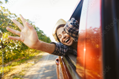 Image of caucasian woman 20s wearing straw hat laughing and waving hand out of the window, while riding in car