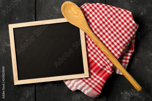 Blank, empty, black chalkboard with wooden cooking spoon and red checkered dish towel flat lay from above on black wooden table