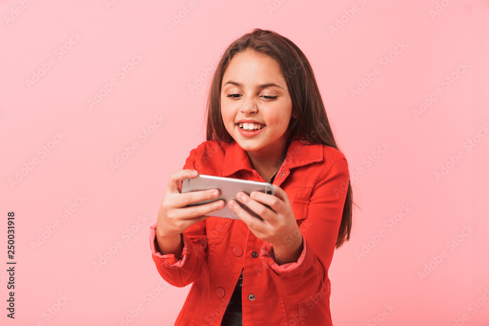 Image of excited teen girl in casual playing video games on cell phone while standing, isolated over red background Image of excited teen girl in casual playing video games on cell phone while standing, isolated over red background