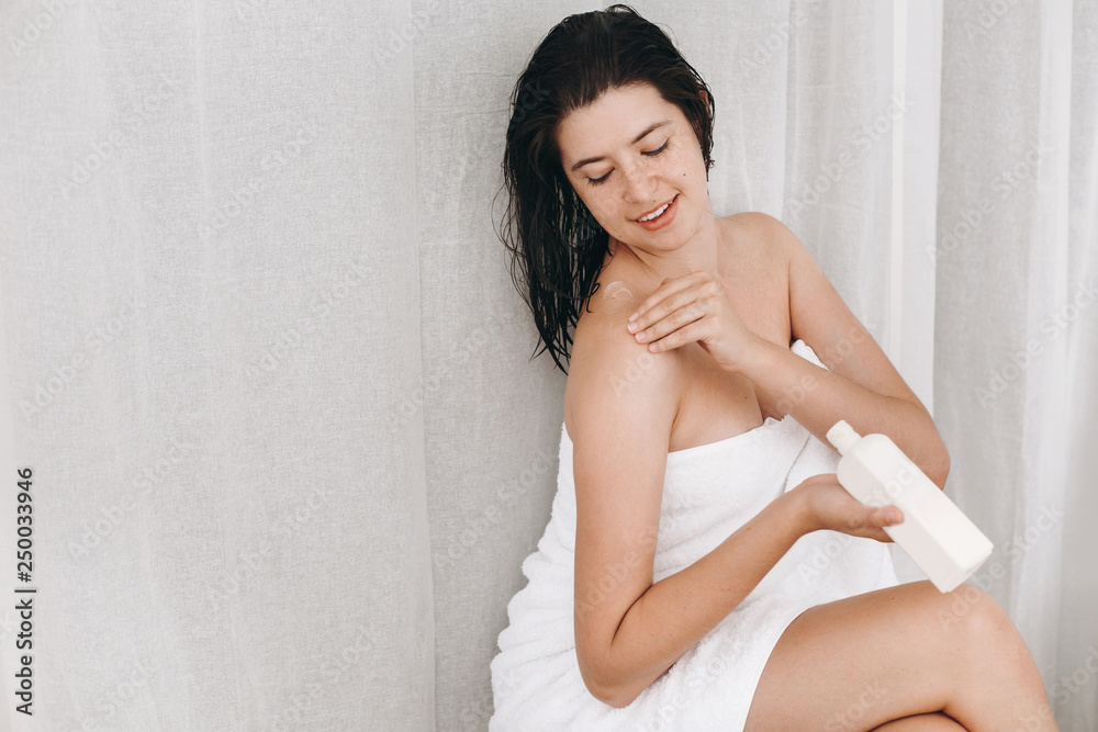 Young happy woman in white towel applying moisturizing cream on shoulder in bathroom. Skin and body care. Hand holding plastic bottle with lotion. Sexy woman relaxing, spa and wellness Young happy woman in white towel applying moisturizing cream on shoulder in bathroom. Skin and body care. Hand holding plastic bottle with lotion. Sexy woman relaxing, spa and wellness