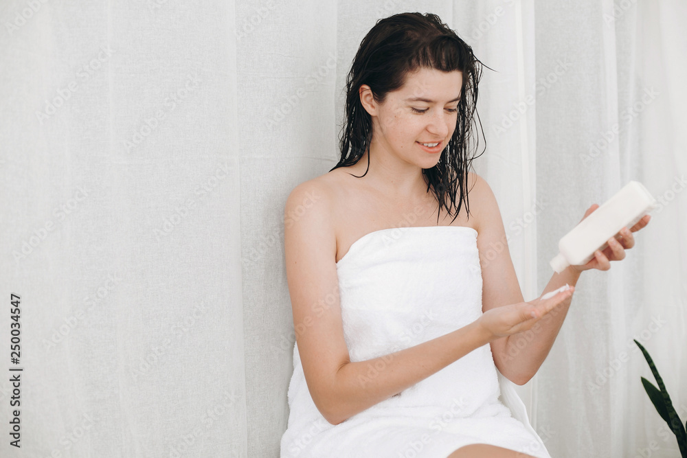 Young woman in white towel applying moisturizing cream on hand in home bathroom with green plants. Skin and body care. Hand holding plastic bottle with lotion. Spa and wellness Young woman in white towel applying moisturizing cream on hand in home bathroom with green plants. Skin and body care. Hand holding plastic bottle with lotion. Spa and wellness