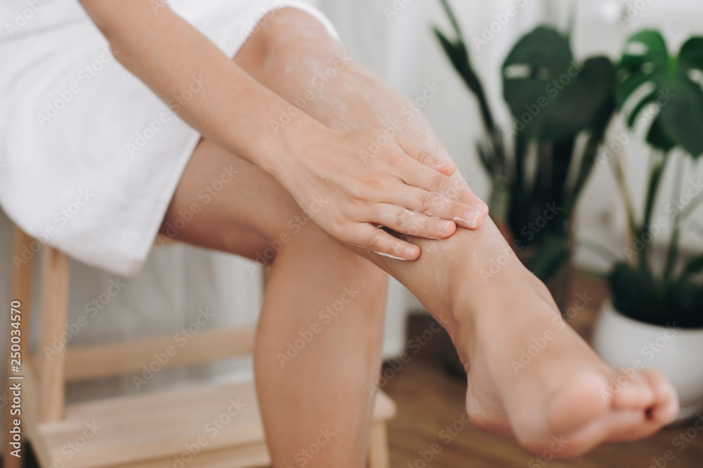 Skin care and wellness concept. Girl hand with moisturizer cream smearing legs for soft skin result. Young woman applying cream on her legs after shaving in bathroom with green plants. Skin care and wellness concept. Girl hand with moisturizer cream smearing legs for soft skin result. Young woman applying cream on her legs after shaving in bathroom with green plants.