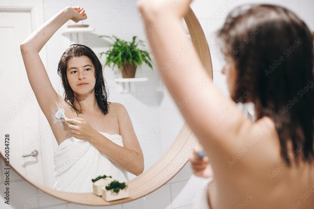 Skin and body care. Hair Removal concept. Woman after shower shaving with razor. Young attractive woman in white towel shaving armpits, looking in mirror in stylish bathroom. Skin and body care. Hair Removal concept. Woman after shower shaving with razor. Young attractive woman in white towel shaving armpits, looking in mirror in stylish bathroom.
