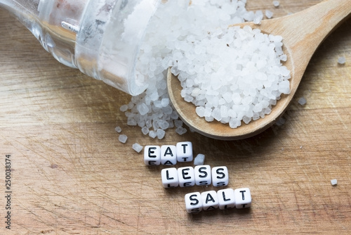 Eat less salt message written near wooden spoon full of granulated salt and shaker on rustic background