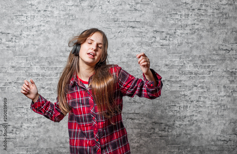 young teenager brunette girl with long hair with headphones listening music and dancing on gray wall background