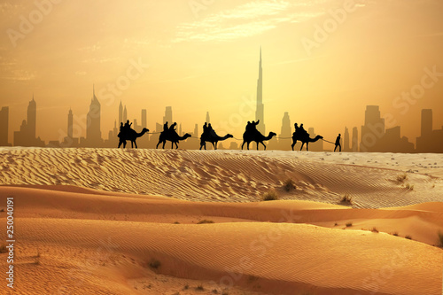 Man and camels in caravan with tourists on sand dunes of Arabian dessert, Dubai skyline seen away 
