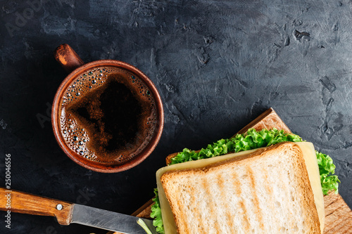 Cup with hot coffee and sandwich with grilled toast, salami sausage, salad lettuce and cheese on dark background, top view