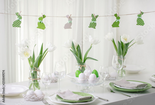 Decor and table setting of the Easter table with white tulips and dishes of green and white color. Easter decor in the form of Easter bunnies  green color with white polka dots.