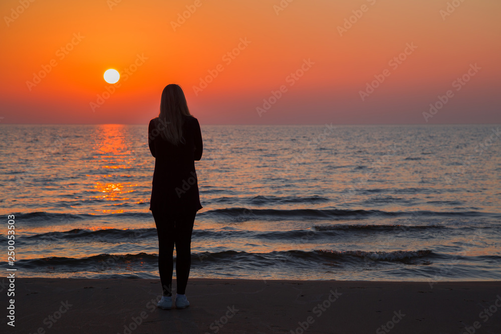 Young adult woman standing alone on sand and staring at small waves of sea and orange sunset. Girl silhouette on beach. Peaceful atmosphere in summer evening. Back view. 