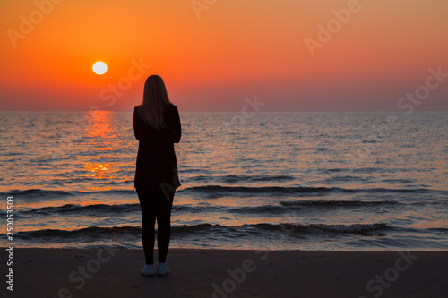 Young adult woman standing alone on sand and staring at small waves of sea and orange sunset. Girl silhouette on beach. Peaceful atmosphere in summer evening. Back view. 