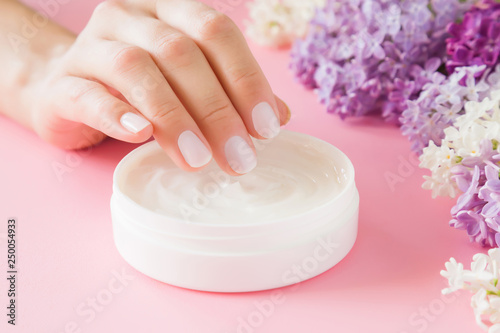 Young  perfect  groomed woman s hands using moisturizing cream. Care about nails and clean  soft  smooth skin. Beautiful branches of fresh  colorful lilac flowers. Front view. Close up. 