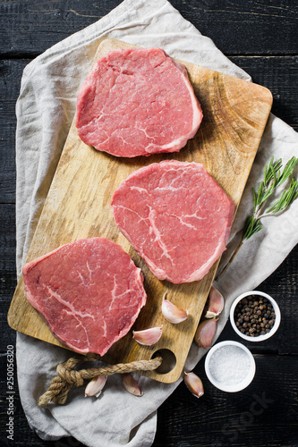 Three raw Beef steaks on a wooden chopping Board, garlic and a sprig of rosemary. Top view, gray background.