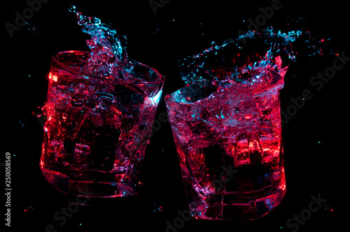 Big splashes of liquid glowing in colorful lights in two rocks glasses clinking in toast isolated on a black background photo