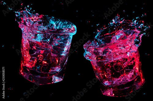 Liquid flying from two full rocks glasses which have just toasted under colorful lights isolated on a black background