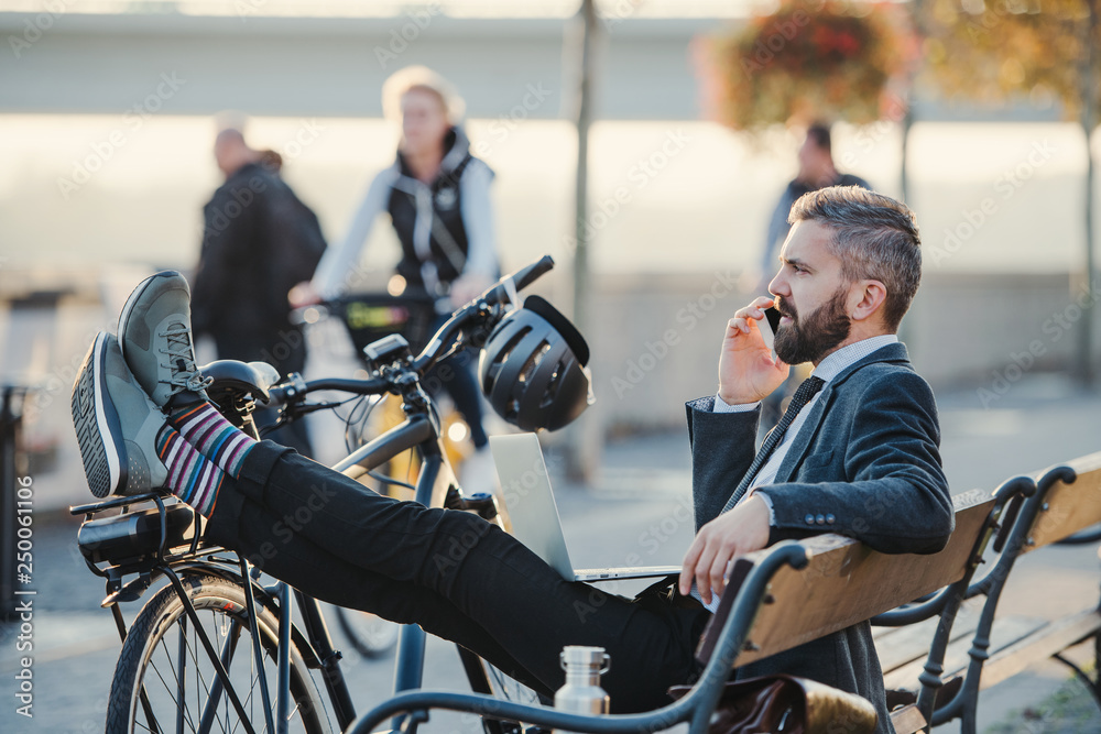 Obraz premium Businessman commuter with bicycle sitting on bench in city, making a phone call.