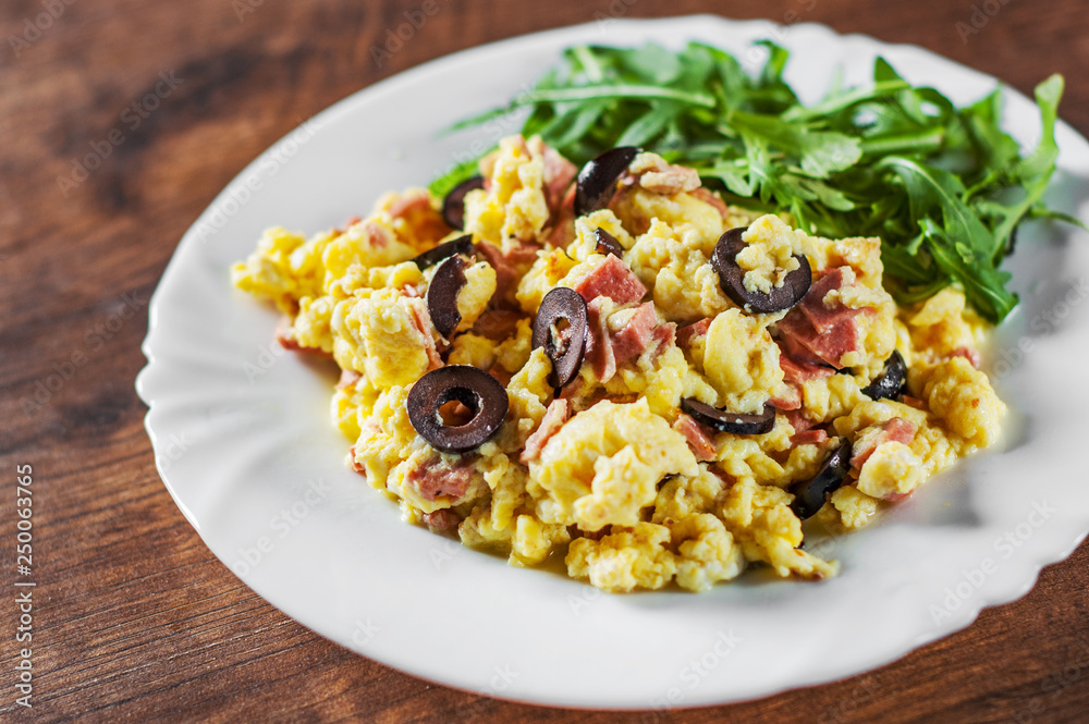 scrambled eggs with ham, olive and arugula salad in white plate on wooden table background scrambled eggs with ham, olive and arugula salad in white plate on wooden table background