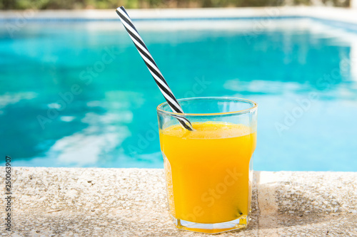 Glass of freshly pressed tropical fruits orange juice with striped straw standing on deck of swimming pool. Bright sunlight. Summer vacation relaxation travel luxury escape. Lifestyle