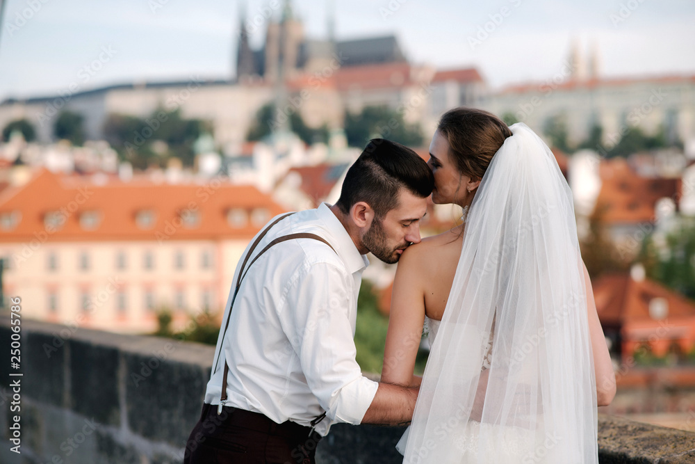 Naklejka premium Back view of young romantic wedding couple standing on the bridge and enjoying beautiful view of the city Prague, Czech Republic. Groom kissing bride