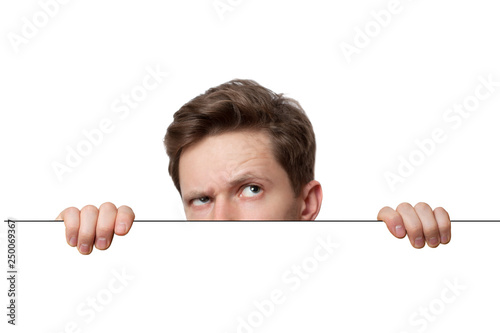 Young man with surprised eyes peeking out from behind billboard paper poster. Man peeking out from the edge and looking at camera isolated on a white background photo